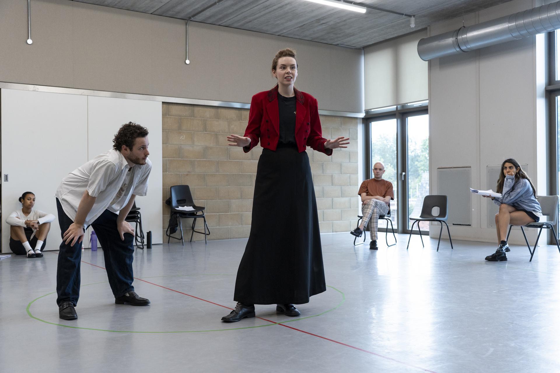 Actors rehearsing a scene in a studio, with a woman in a red jacket gesturing while speaking and others watching from chairs around a taped floor circle